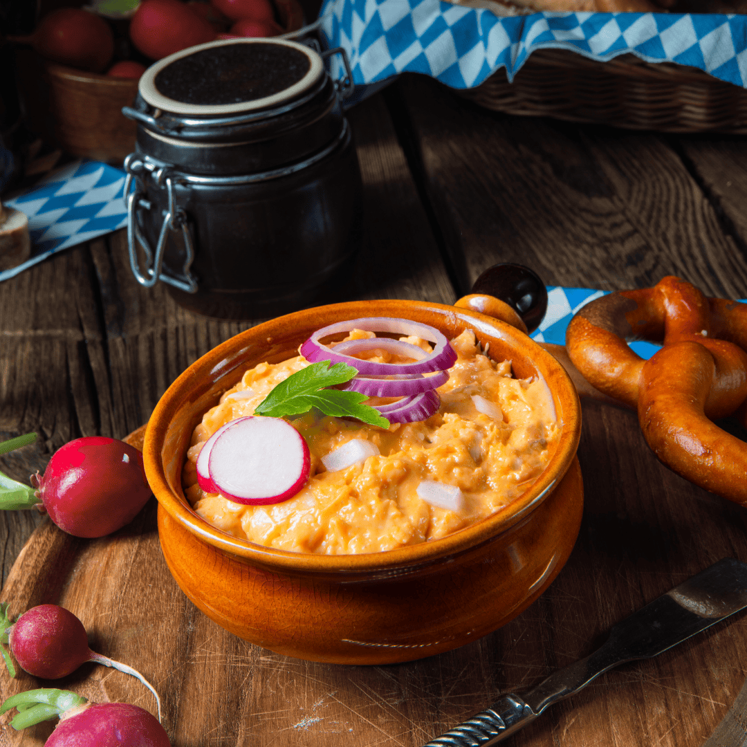 Traditional Bavarian Obazda cheese spread in a rustic ceramic bowl, garnished with red onion rings, radish slices, and parsley, served with pretzels on a wooden table.