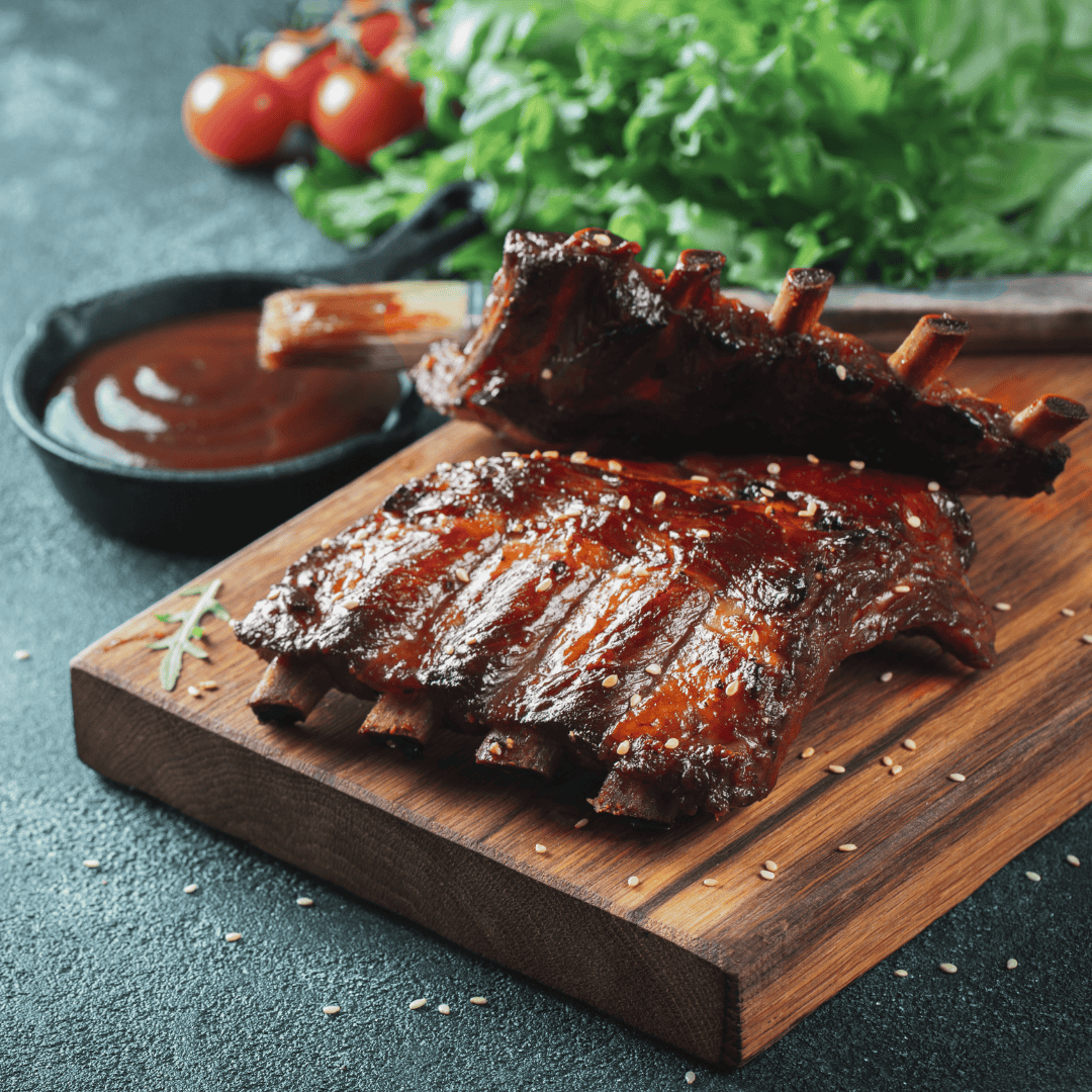 Close-up of glazed BBQ spare ribs sprinkled with sesame seeds on a wooden board, served with a bowl of barbecue sauce, fresh lettuce, and cherry tomatoes in the background.