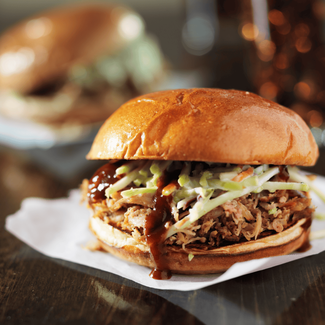 Close-up of a pulled pork sandwich topped with coleslaw and drizzled with BBQ sauce in a soft brioche bun, styled with an Iron Maiden–themed background