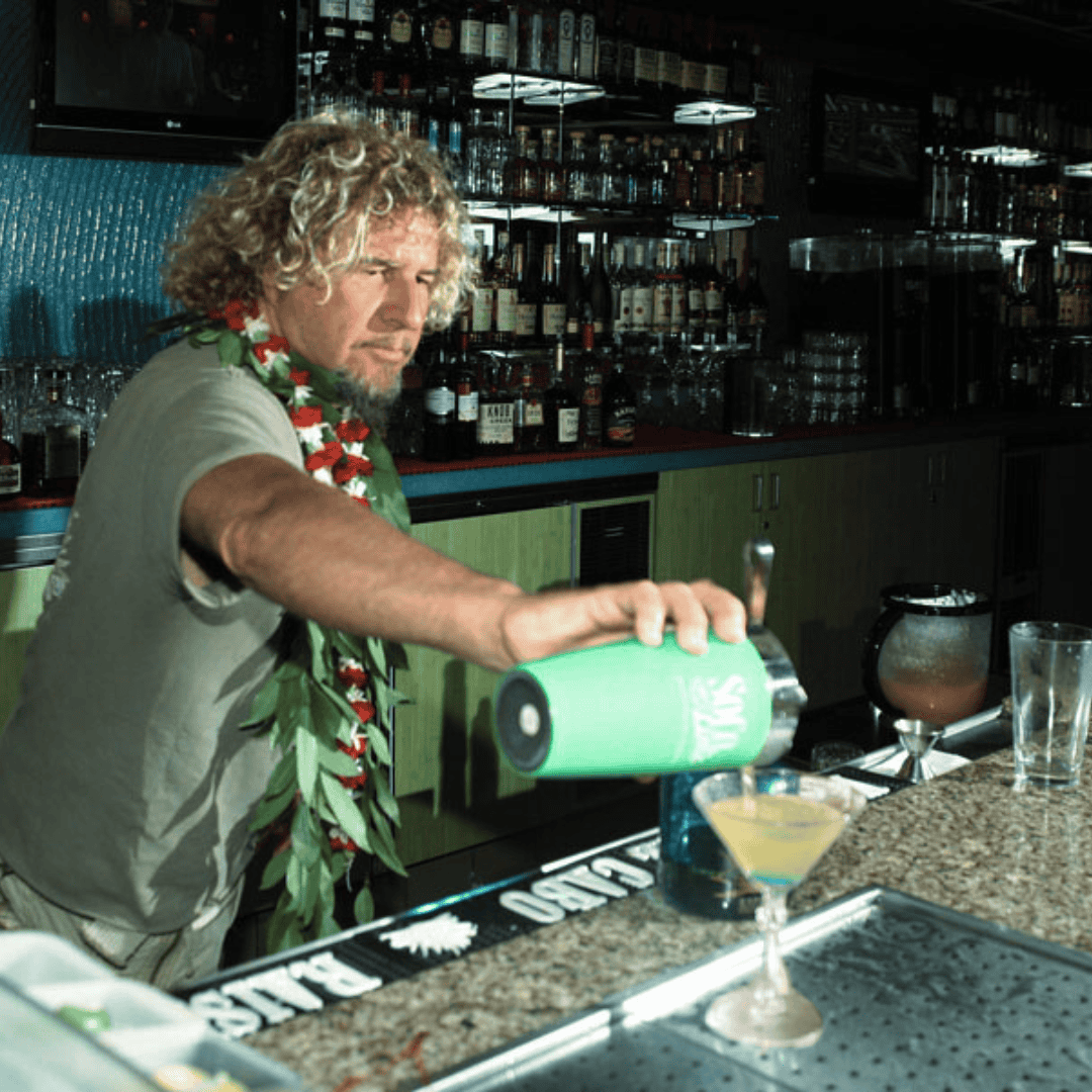 Sammy Hagar wearing a Hawaiian lei pours a margarita from a cocktail shaker at a bar, with liquor bottles in the background.