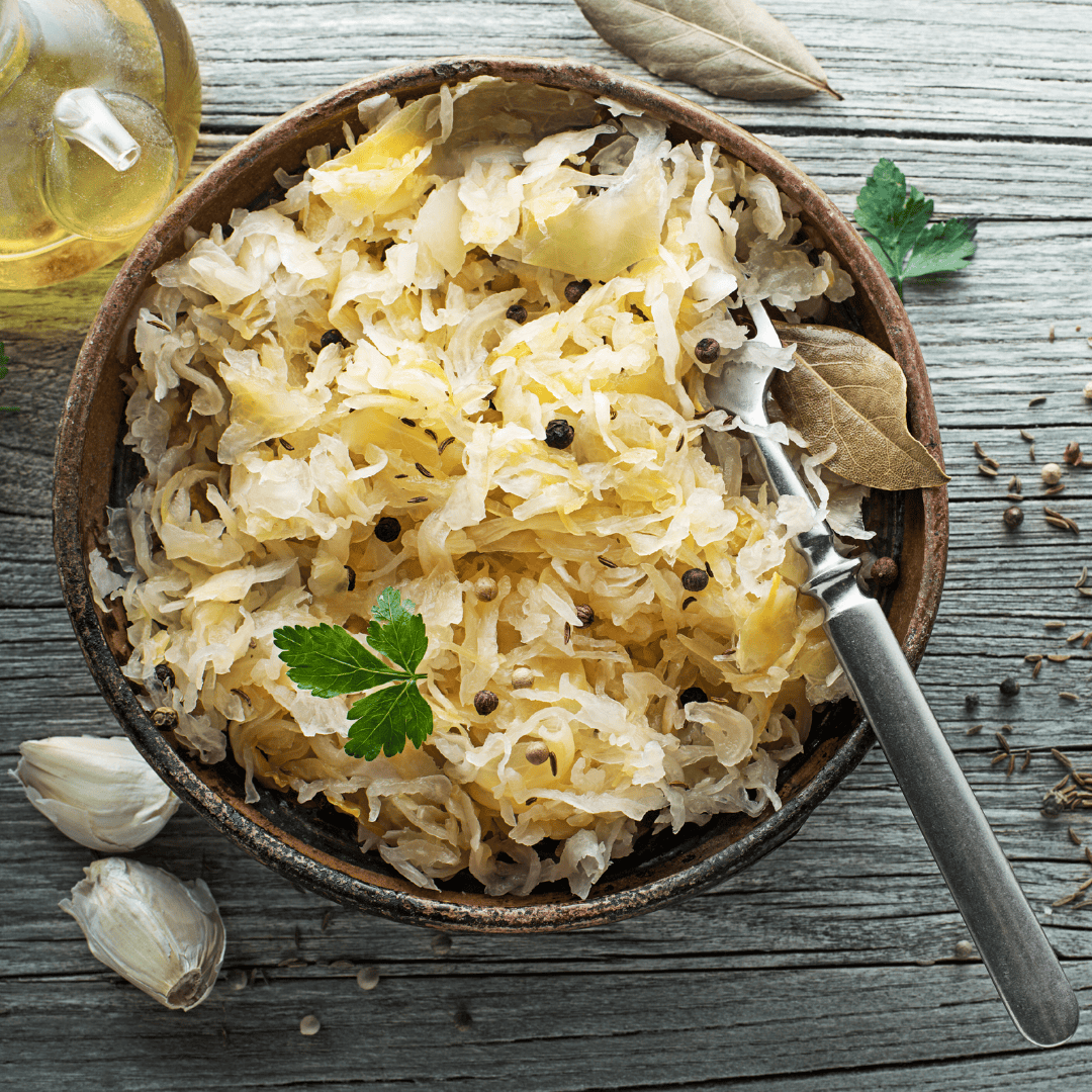 Bowl of homemade sauerkraut with peppercorns, bay leaves, and fresh parsley garnish, served on a rustic wooden table with garlic cloves and caraway seeds.