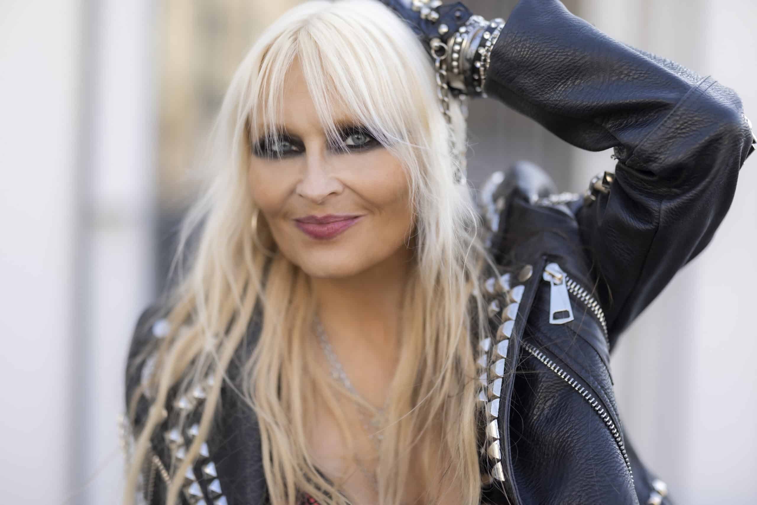 Portrait of Doro Pesch wearing a black leather jacket with metal studs and zippers, smiling confidently with one arm raised, showcasing her signature heavy metal style.