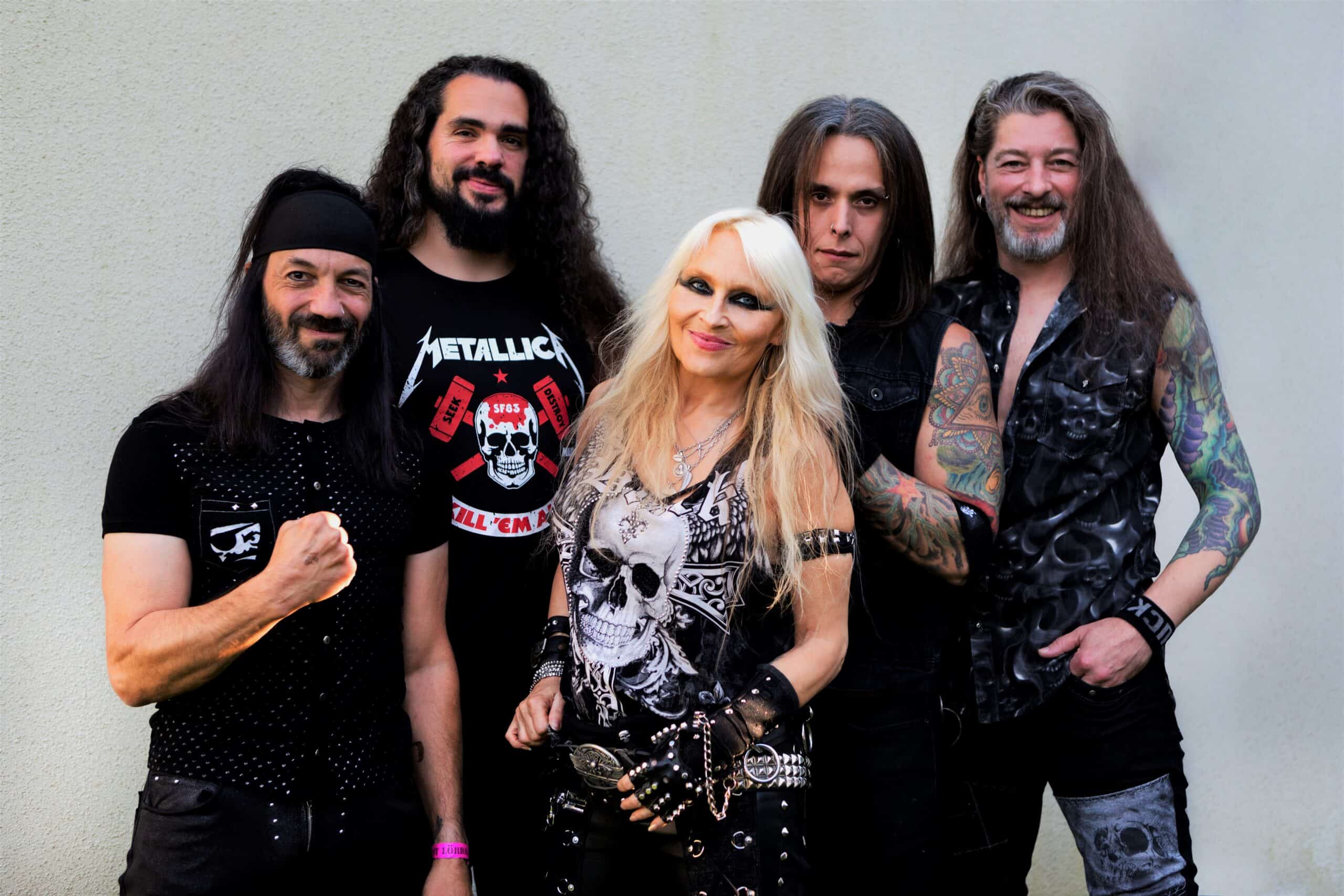 Doro Pesch standing with her band members, all dressed in heavy metal attire with skull motifs, leather, tattoos, and long hair, posing confidently against a light wall.